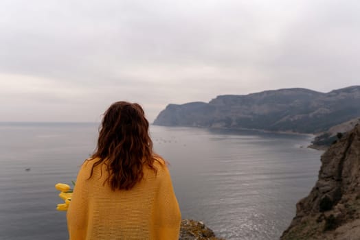 Rear view of a woman with long hair against a background of mountains and sea. Holding a bouquet of yellow tulips in her hands, wearing a yellow sweater.