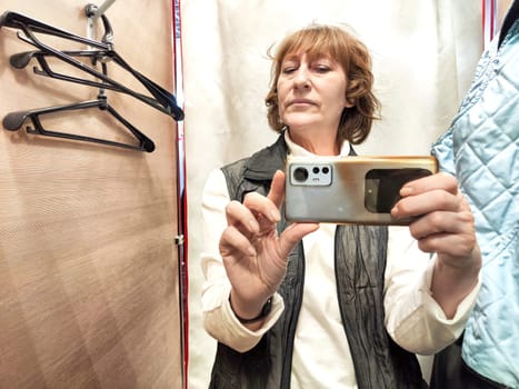 A middle-aged blonde woman in a cramped fitting room of a store with a bunch of clothes and hangers takes selfie in the mirror