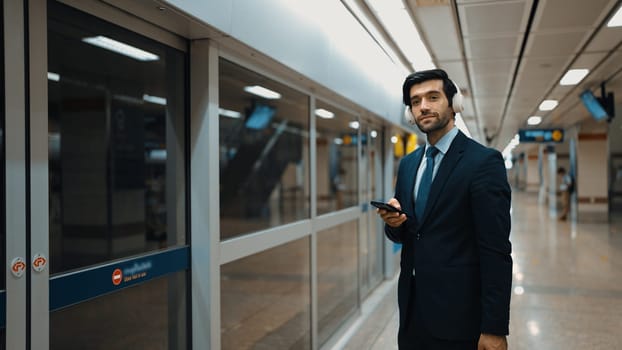 Project manager wearing headphone at train station while holding mobile phone for choosing song. Smart business man listening relaxing music while waiting for train with blurred background. Exultant.