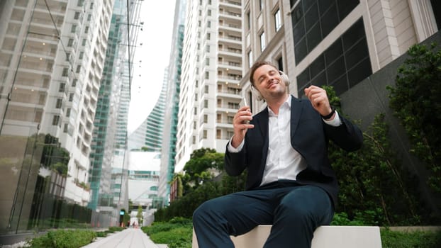 Caucasian businessman listening relaxing music while hold mobile phone. Skilled happy executive manager open music and move to song in lively mood while sitting at green urban city or park. Urbane.