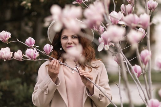 Woman magnolia flowers, surrounded by blossoming trees, hair down, white hat, wearing a light coat. Captured during spring, showcasing natural beauty and seasonal change