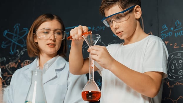 Close up of caucasian boy mixing chemical liquid while teacher giving advice. Instructor wearing lab suit looking for diverse student at table with beaker filled with colored solution. Erudition.