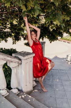 woman in a red silk dress and a bandage on her head smiles against the background of the leaves of a tree. She is leaning on the coop and looking into the camera. Vertical photo