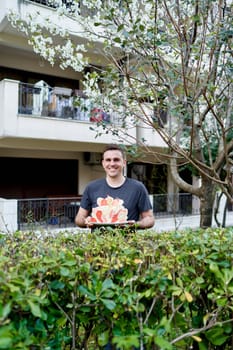 Smiling young man with colorful birthday cake on plate standing near green bushes in garden. High quality photo