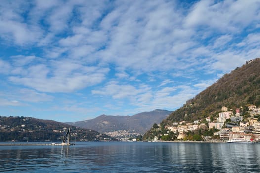 Beautiful view of the lake of Como against Alps mountains and blue cloudy sky background on a sunny winter day in the province of Milan in Lombardy, Italy. Lago di Como. Wonderful travel destinations
