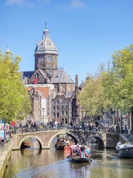 Amsterdam Netherlands 21 April 2024 A boat with tourist peacefully glides down a canal river beside a towering, majestic building, creating a striking contrast between natural and urban landscapes.