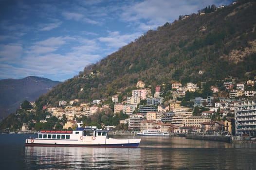 A boat on the lake of Como against blue sky and Italian Alps mountains background. Clear water of the lake reflecting floating boat and houses on the waterfront. Nature background, Tourism and travel.
