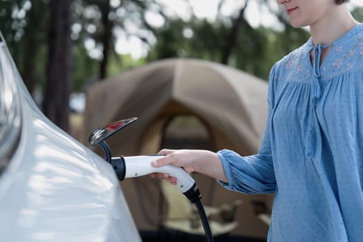 Holiday camping vacation traveling to nature countryside with electric car, woman recharge EV car from charging station on campsite. Natural travel and eco car for sustainable environment. Perpetual