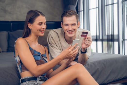 Young couple sit in the home bedroom using online payment app and digital wallet on smartphone to pay with credit card. E commerce shopping and modern purchasing via mobile internet. Unveiling