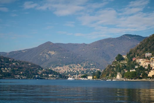 Beautiful view of the lake of Como against Alpine mountains background. Clear water of the lake reflecting floating boats and houses on the waterfront. Italy. Nature background, Tourism and travel.