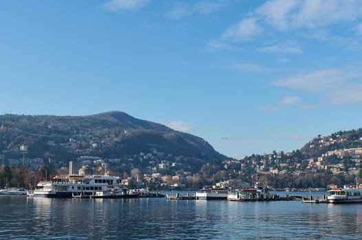 Beautiful view of floating boats and ships reflecting in the clean clear water of the lake of Como, against Italian Alps and blue sky background on a sunny cold winter day. Tourism and travel concept