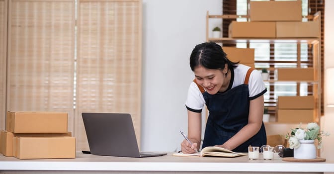 Young woman, small business owner selling products online, accepting online product orders via laptop, sits in a room with boxes of products..