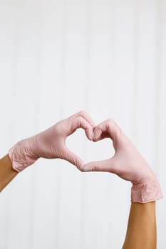 Hands with gloves making heart shape symbolize love or support to medical team. Person in pink latex gloves showing heart gesture against white background, closeup on hands.