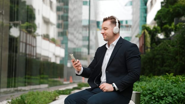 Caucasian businessman listening relaxing music while hold mobile phone. Skilled happy executive manager open music and move to song in lively mood while sitting at green urban city or park. Urbane.