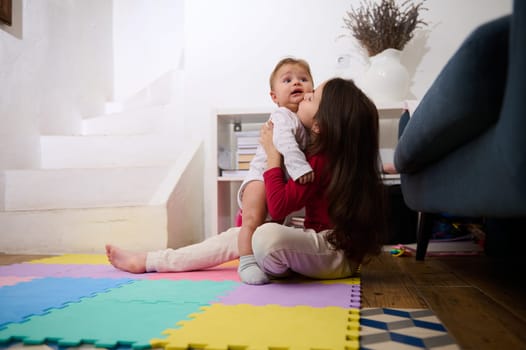 Adorable little child girl sitting on colorful puzzle carpet, holding and kissing her little brother, playing with a cute baby boy in light interior of a cozy rural house. Family relationships concept