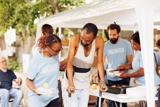 Multiethnic charity group handing out free food and nourishments to less fortunate and disabled. Portrait of black woman with blue t-shirt assisting the poor, needy african american man on crutches.