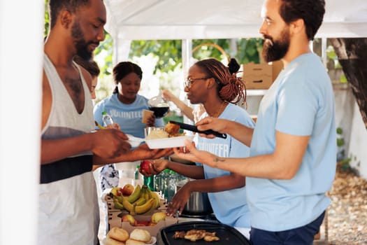 Young African-American and Caucasian voluntary individuals handing out free food and necessities to underprivileged. At homeless shelter, friendly volunteers give hunger relief and humanitarian aid.