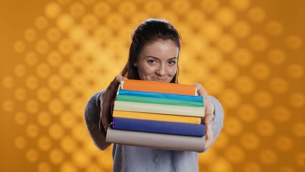 Portrait of friendly woman offering stack of textbooks useful for school exam, isolated over studio background. Merry person giving pile of books, recommending them for university assessment, camera B