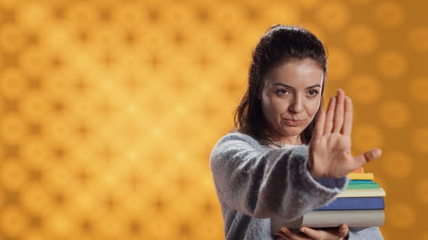 Portrait of stern woman holding stack of books doing stop sign gesturing, studio background. Student with pile of textbooks in arms used for academic learning doing halt hand gesture, camera A