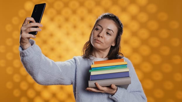 Smiling woman holding pile of books, enjoying reading hobby, taking selfies with cellphone. Cheerful lady with stack of novels in arms doing pictures with mobile phone, studio background, camera B