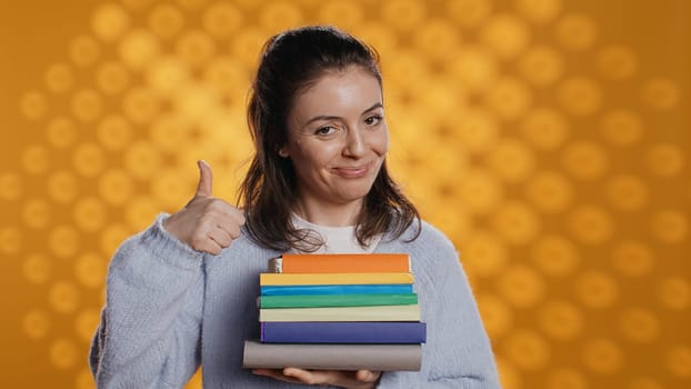 Portrait of happy woman with stack of books in hands showing thumbs up, studio background. Joyous bookworm holding pile of novels, feeling upbeat, doing positive hand gesturing, camera B