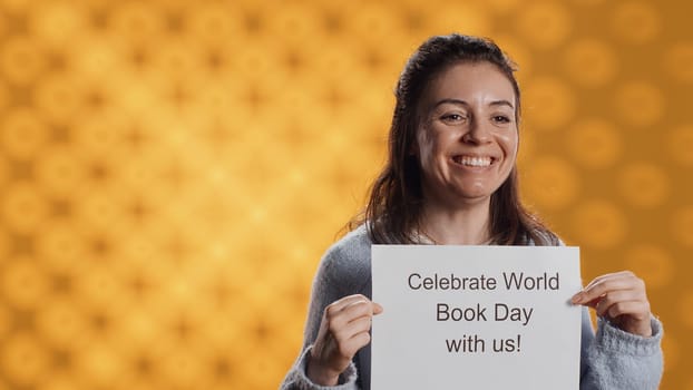 Portrait of happy woman holding placard with world book day message written on it, isolated over studio background. Geek promoting importance of reading during 23th April global event, camera A