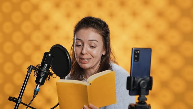 Media star filming vlog at work, reading book to record audiobook, studio background. Voice actor doing content creation while producing digital recording on novel using microphone, camera A