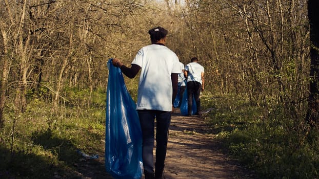 African american girl collecting rubbish in a trash bag using tongs, recycling plastic waste and picking up garbage in a forest. Young woman activist clearing the woods from junk. Camera B.
