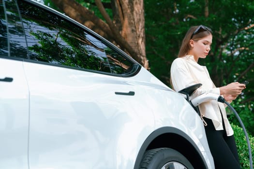 Young woman use smartphone to pay for electricity at public EV car charging station green city park. Modern environmental and sustainable urban lifestyle with EV vehicle. Expedient