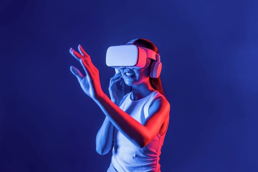 Smart female standing with surrounded by cyberpunk neon light wear VR headset connecting metaverse, futuristic cyberspace community technology. Woman using hand touching virtual object. Hallucination.