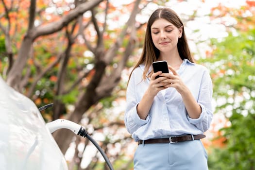 Young woman using smartphone online banking application to pay for electric car battery charging from EV charging station during autumn vacation holiday trip at national park or autumnal forest. Exalt