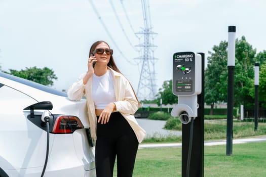 Young woman recharging EV car battery while talk on phone at charging station connected to electrical power grid tower facility as electrical industry for eco friendly vehicle utilization. Expedient