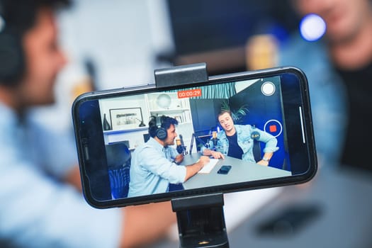 Host channel of influencers talking in broadcast streaming online wearing headsets with script on social media with smartphone recording, greeting listeners at modern studio work place. Sellable.