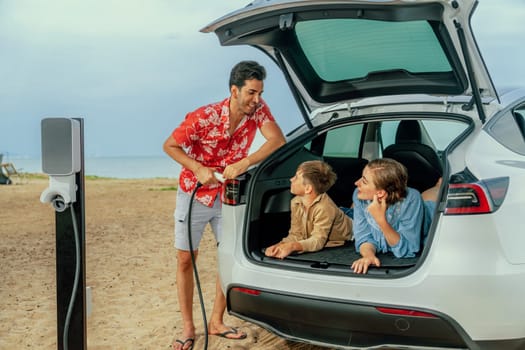 Family vacation trip traveling by the beach with electric car, lovely family sit on the trunk, charging EV car battery with green and sustainable energy. Family travel and eco-friendly car. Perpetual