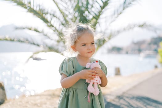 Little girl hugs a toy pink mouse while standing on the seashore near a green palm tree. High quality photo