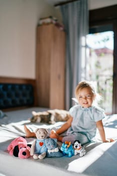 Teddy bear and soft toys sit in a row on the bed against the background of a little smiling girl. High quality photo