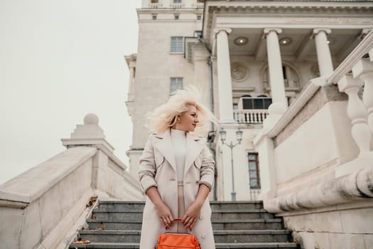 A woman wearing a white coat and blonde hair stands on a set of stairs in front of a large building. She is holding a red purse in her hand. Concept of elegance and sophistication