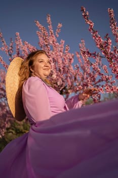 Woman blooming peach orchard. Against the backdrop of a picturesque peach orchard, a woman in a long pink dress and hat enjoys a peaceful walk in the park, surrounded by the beauty of nature