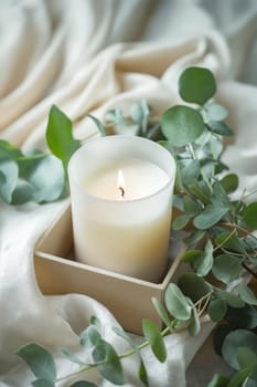 A candle is lit in a glass container on a table with green leaves. The candle is surrounded by a box and a white cloth. Generative AI
