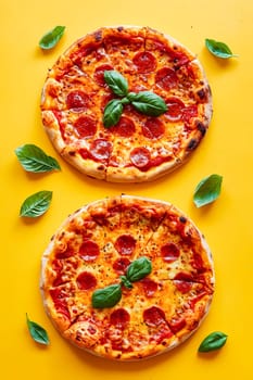 Two pizzas with pepperoni and basil on top of a yellow background. The pizzas are placed next to each other and surrounded by green leaves. Generative AI