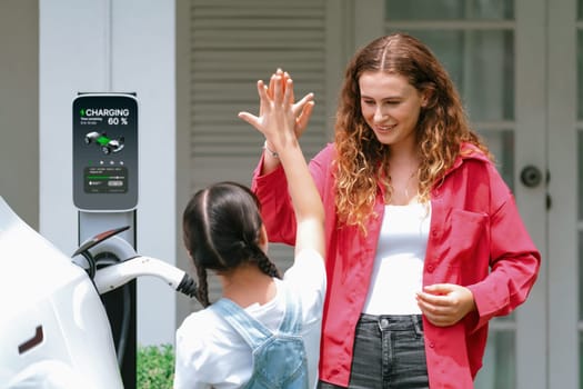 Happy little young girl learn about eco-friendly and energy sustainability as she help her mother recharge electric vehicle from home EV charging station. EV car and modern family concept. Synchronos