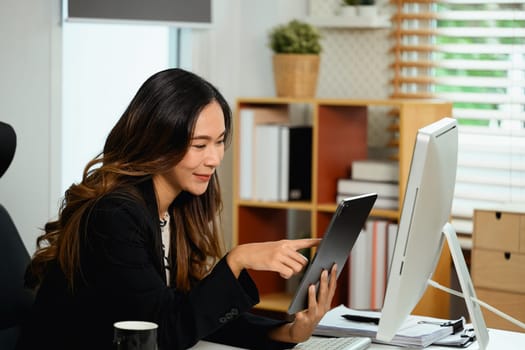 Cheerful young businesswoman sitting at desk and using digital tablet.