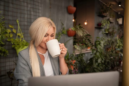 A blonde woman is sitting at a table with a laptop and a white coffee cup. She is drinking coffee while working on her laptop