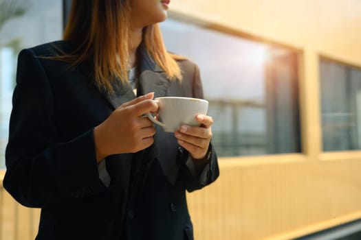 Businesswoman in black suit looking away while standing with coffee cup in modern office.