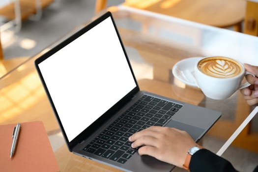 Cropped shot of young woman holding a cup of coffee with latte art and using laptop at cafe.