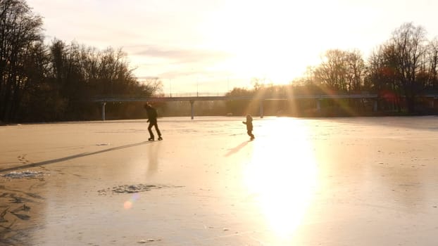 Elder Brother playing with young brother on the frozen river in sunset lightning. Winter landscape games skating on the ice concept.
