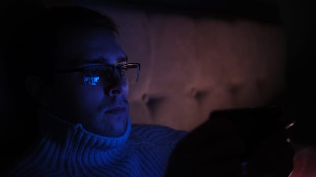 Lonely man in glasses sitting on bed in bedroom and using phone at night. Face illuminated from smartphone display. Surfing social network before sleep.