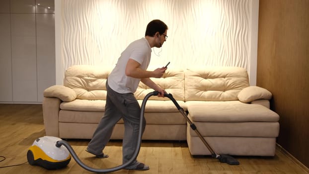 Young caucasian man in white and gray clothes having fun and dancing while vacuuming at home with biege sofa on background. Man in earphones with phone in hand have dancing with vacuum cleaner.