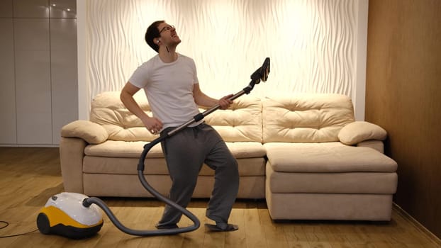 Young caucasian man in white and gray clothes having fun and dancing while vacuuming at home with biege sofa on background. Man in earphones have dancing with vacuum cleaner.