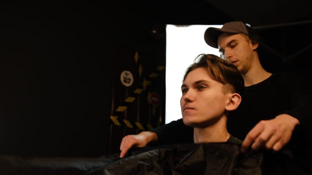 Caucasian man in shirt sitting on the chair while barber closing his client with black peignoir in salon. Hairdresser service in a modern barbershop in a dark key lightning with warm light side view.